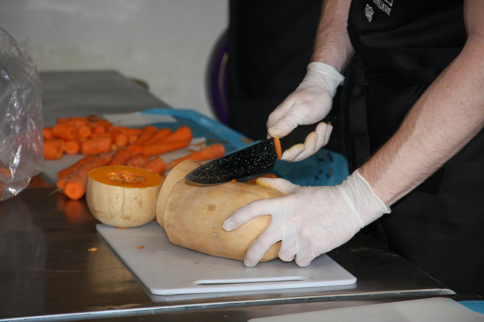 A kitchen volunteer in white gloves slicing a butternut on a chopping board, beside a pile of chopped carrots.
