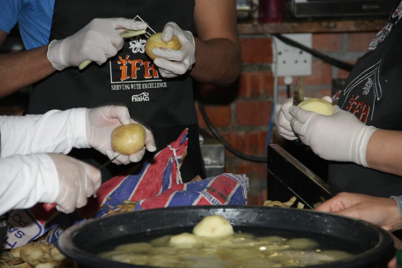 Four volunteers in black Ihsan Family Homes aprons and white gloves peeling potatoes together over a large pot, in the kitchen.